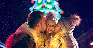 Un padre y una madre dan un beso a su hija. De fondo un arbol de navidad iluminado.